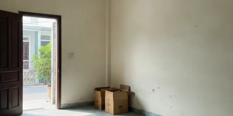 Phòng khách trống của một căn nhà phố, tường trơn và vài thùng giấy xếp ở góc, cửa mở ra ngoài sáng