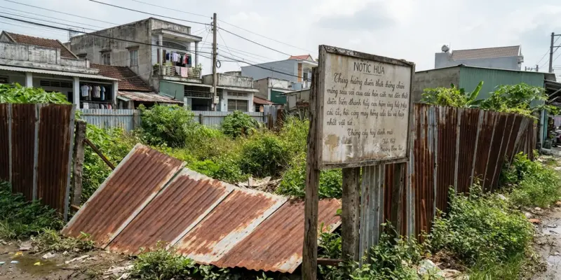 Thửa đất trống quây tôn cũ, cỏ mọc cao, có tấm biển thông báo bạc màu đóng trên cọc
